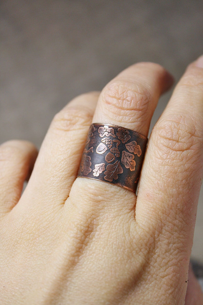 Puede incluir: Un anillo ancho de cobre con un dise&ntilde;o grabado de hojas de roble y bellotas. El anillo se lleva en un dedo.