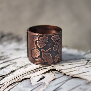 Puede incluir: Un anillo de cobre con un dise&ntilde;o grabado de hojas de roble y bellotas. El anillo est&aacute; sobre un fondo de corteza de abedul blanca.