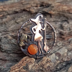 May include: A copper ring with a silhouette of a witch holding a broom and a small orange gemstone. The ring is set on a piece of wood.