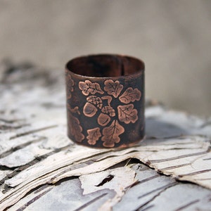 Puede incluir: Un anillo de cobre con un dise&ntilde;o grabado de hojas de roble y bellotas. El anillo est&aacute; sobre un trozo de corteza de abedul.