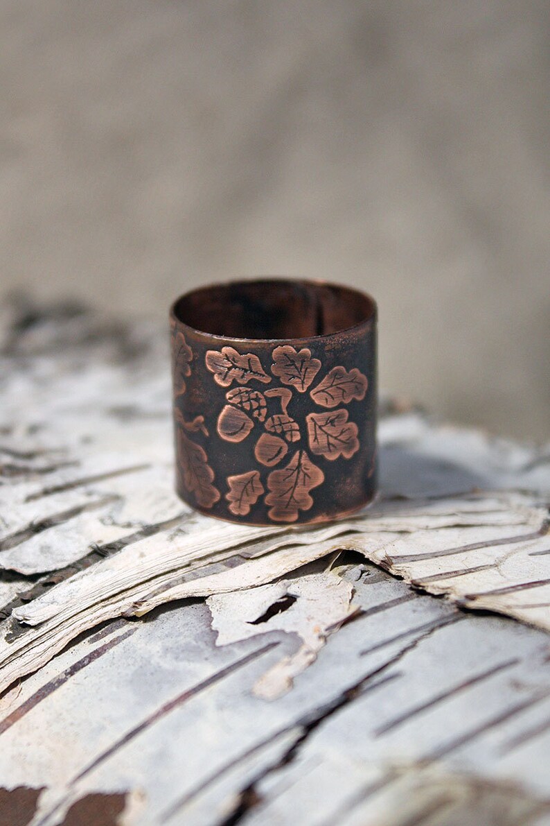 Puede incluir: Un anillo de cobre con un dise&ntilde;o grabado de hojas de roble y bellotas. El anillo est&aacute; sobre un trozo de corteza de abedul.