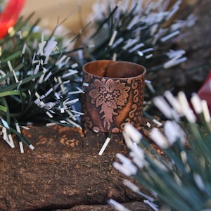 Puede incluir: Un anillo de cobre con un diseño floral. El anillo está sentado en un trozo de madera con oropel verde y blanco.