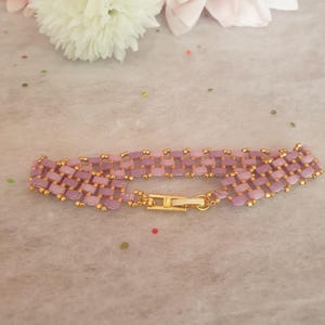 Peut inclure: Un bracelet de perles tissées, composé de perles lavande et dorées. Le bracelet est doté d'un fermoir doré et est présenté sur une surface douce et texturée. Le bracelet mesure environ 18 cm de long.