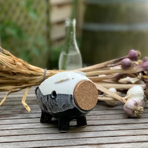 May include: A ceramic pig-shaped bottle stopper with a cork. The pig is white and black with a textured finish. The stopper is on a wooden table with a blurred background of a glass bottle, dried plants, and garlic.