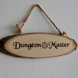 Może przedstawiać: Drewniany znak z napisem "Dungeon Master" i symbolem dwudziestościennej kostki do gry.