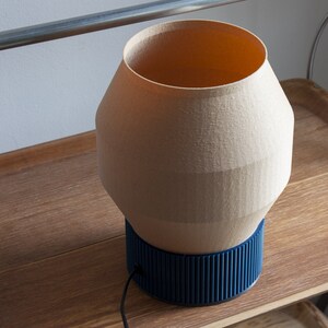 May include: A beige ceramic table lamp with a blue ribbed base. The lamp is sitting on a wooden surface.