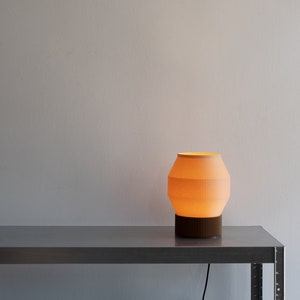 May include: A warm, orange-toned table lamp with a white, ribbed shade sits on a silver metal table. The lamp is turned on and casting a soft glow.