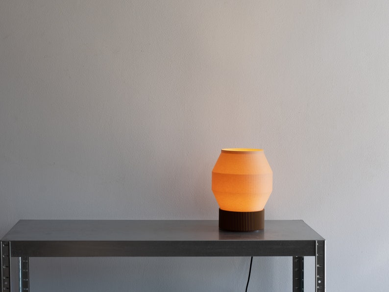 May include: A warm, orange-toned table lamp with a white, ribbed shade sits on a silver metal table. The lamp is turned on and casting a soft glow.