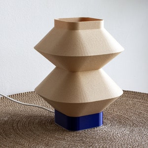 Może przedstawiać: Nowoczesna lampa stołowa o geometrycznym, warstwowym designie. Lampa posiada beżowy, teksturowany klosz i kwadratową, niebieską podstawę. Lampa jest podłączona białym przewodem i stoi na brązowej, okrągłej macie.
