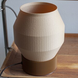 May include: A beige lamp with a brown base. The lamp has a unique geometric shape and a textured fabric shade.