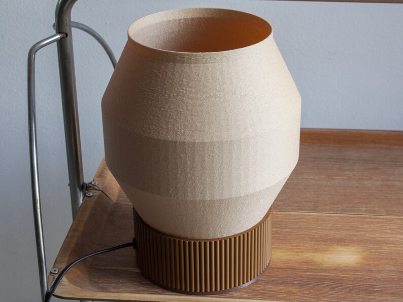 May include: A beige lamp with a brown base. The lamp has a unique geometric shape and a textured fabric shade.