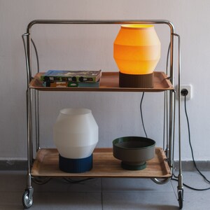 May include: Two lamps on a metal rolling cart with wooden shelves. The top lamp has a yellow shade and the bottom lamp has a white shade. A book is on the top shelf.