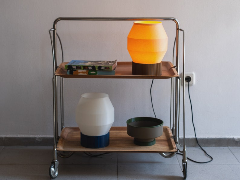 May include: Two lamps on a metal rolling cart with wooden shelves. The top lamp has a yellow shade and the bottom lamp has a white shade. A book is on the top shelf.