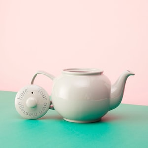 May include: A light grey ceramic teapot with a lid that says "Your own message here". The teapot is sitting on a teal surface.