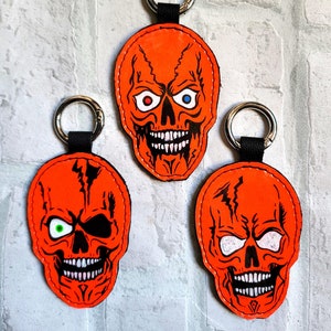 May include: Three orange leather keychains featuring skull designs. Each skull has black eyes with different coloured pupils: red, green, and white. The skulls have black outlines and white teeth.