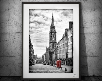 May include: A black and white photograph of a street in Edinburgh, Scotland. The street is lined with buildings, and a tall church spire is visible in the distance. A red telephone booth is in the foreground.