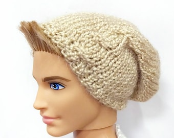 May include: A beige knitted beanie hat. The hat is made of a chunky yarn and has a slouchy fit. The hat is shown on a doll with blonde hair and blue eyes. The doll is wearing a white and blue sweater.