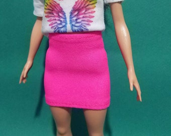 May include: A white t-shirt with short sleeves and a rainbow wing design. The shirt has the text "FLY to YOUR dream" printed on it. The doll is wearing a pink skirt.