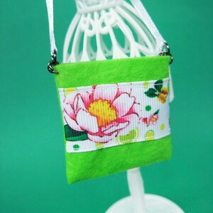Puede incluir: Una peque&ntilde;a bolsa de fieltro verde con estampado floral blanco y rosa. La bolsa tiene una correa blanca y cierres plateados.