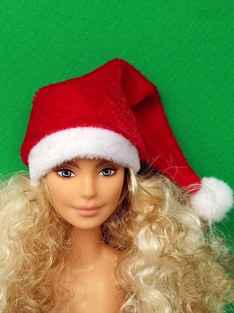 Puede incluir: Una mu&ntilde;eca rubia con ojos azules que lleva un gorro de Pap&aacute; Noel rojo con ribete blanco. La mu&ntilde;eca tiene el pelo rizado y no lleva ropa.