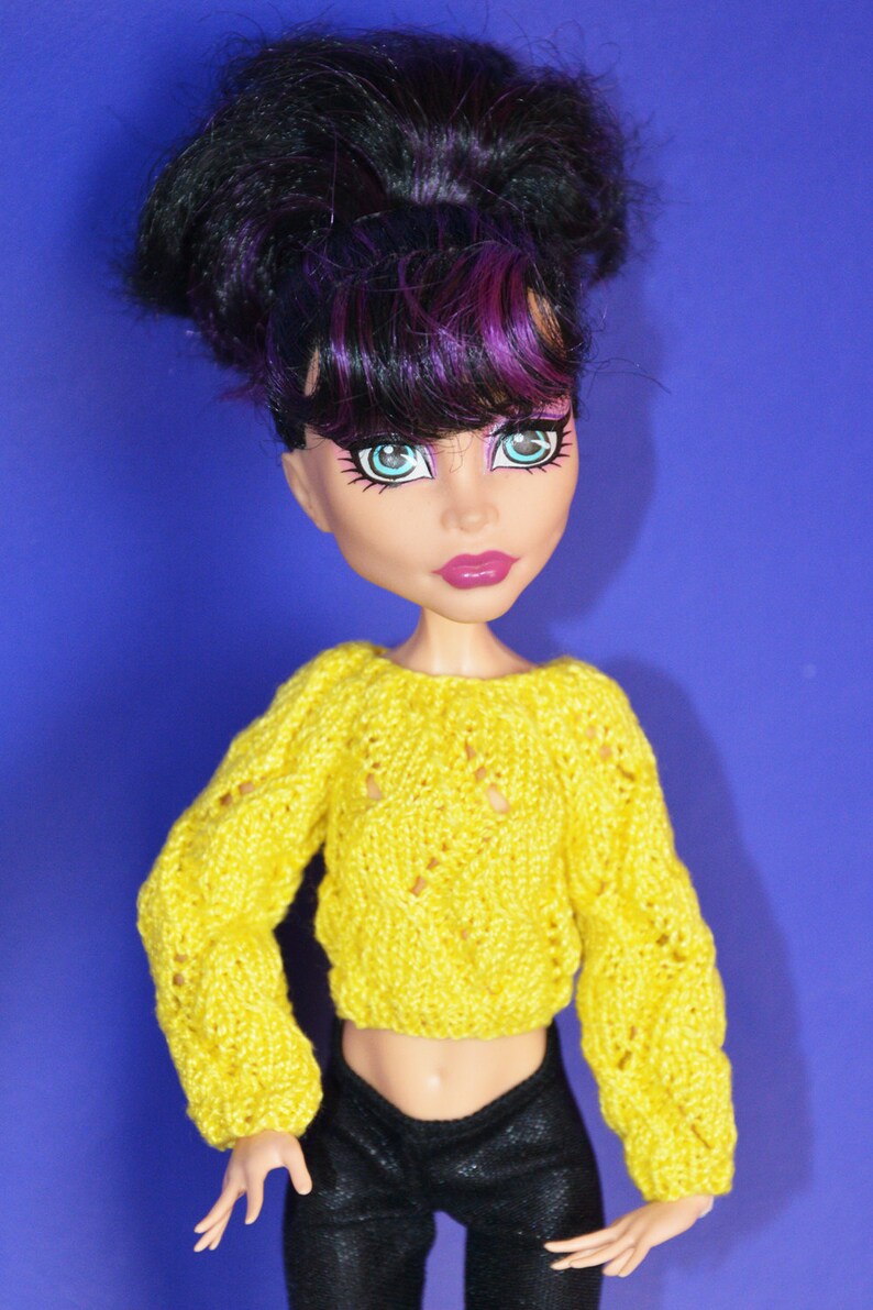 May include: A doll with black and purple hair wearing a yellow knitted sweater and black pants.