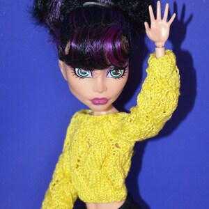 May include: A doll with black and purple hair wearing a yellow knitted sweater and black pants. The doll is holding up one hand and has a bright blue eye.
