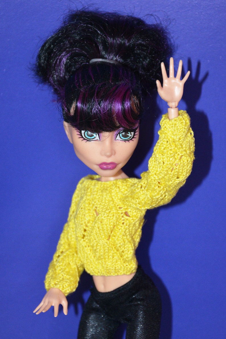 May include: A doll with black and purple hair wearing a yellow knitted sweater and black pants. The doll is holding up one hand and has a bright blue eye.