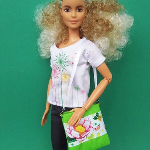 Puede incluir: Una mu&ntilde;eca con el pelo rubio rizado que lleva una camiseta blanca con un dise&ntilde;o de fuegos artificiales y unos vaqueros negros. La mu&ntilde;eca lleva un bolso bandolera floral verde y blanco.