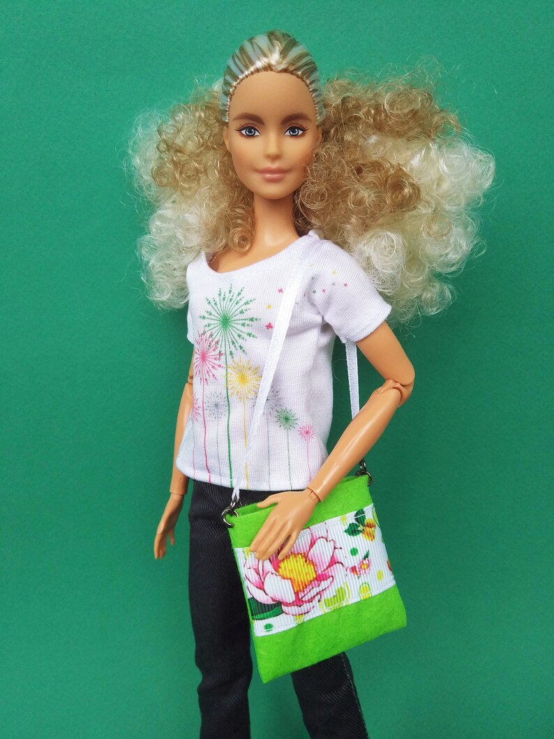 Puede incluir: Una mu&ntilde;eca con el pelo rubio rizado que lleva una camiseta blanca con un dise&ntilde;o de fuegos artificiales y unos vaqueros negros. La mu&ntilde;eca lleva un bolso bandolera floral verde y blanco.