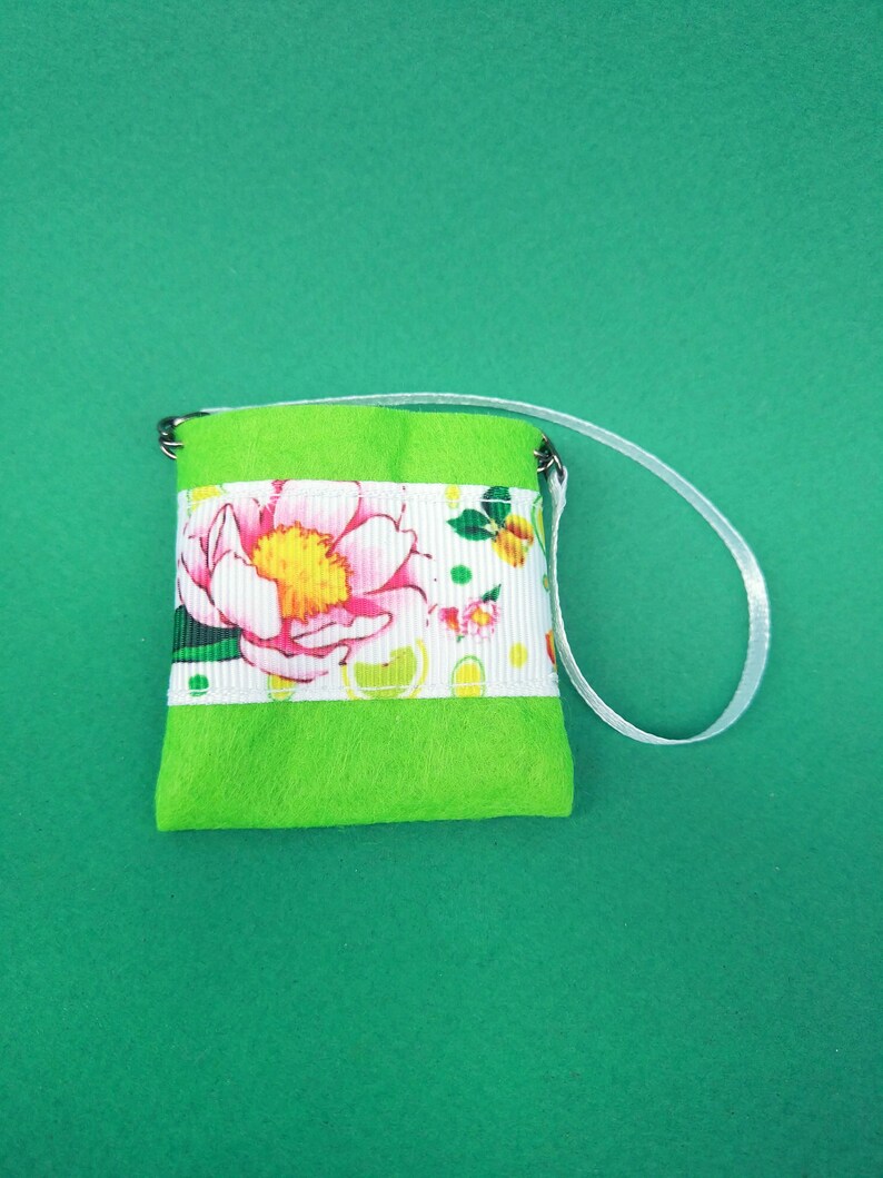 Puede incluir: Una peque&ntilde;a bolsa de fieltro verde con una cinta floral blanca y un asa de cinta blanca.