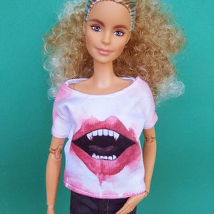 May include: A doll with blonde curly hair wearing a white t-shirt with a red and black vampire mouth graphic. The doll is wearing black trousers and has blue eyes.