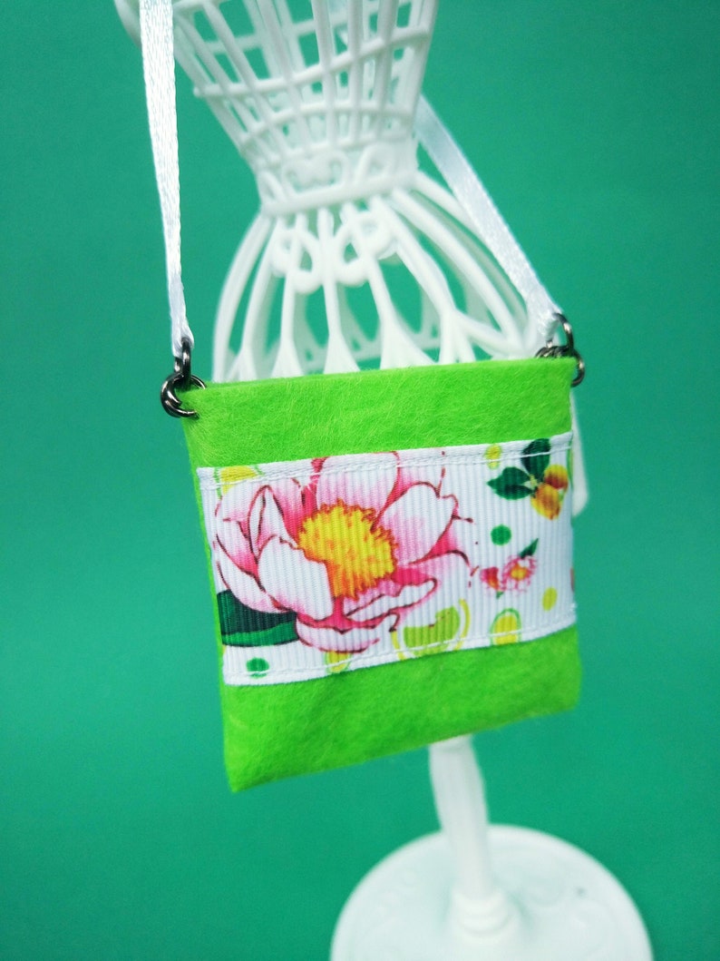 Puede incluir: Una peque&ntilde;a bolsa de fieltro verde con estampado floral blanco y rosa. La bolsa tiene una correa blanca y cierres plateados.