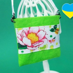 Puede incluir: Un peque&ntilde;o bolso de fieltro verde con un ribete de cinta floral blanca y rosa. El bolso tiene una correa de cadena plateada.