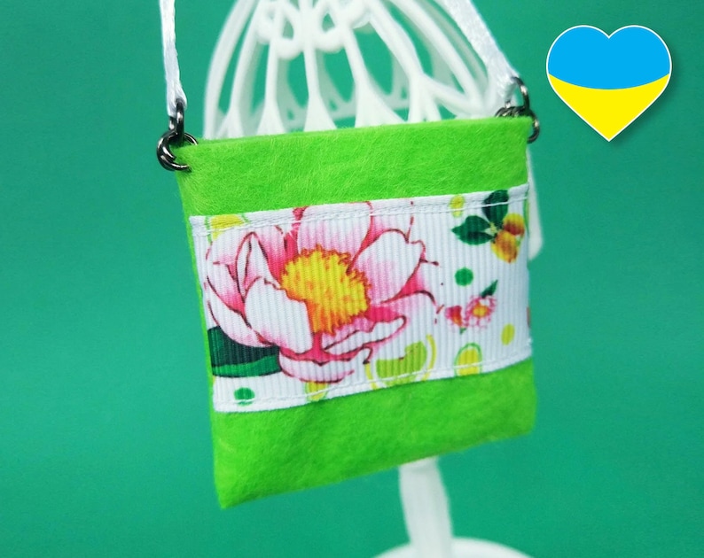Puede incluir: Un peque&ntilde;o bolso de fieltro verde con un ribete de cinta floral blanca y rosa. El bolso tiene una correa de cadena plateada.
