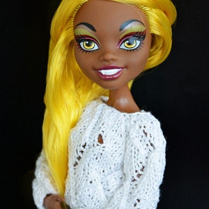 May include: A doll with yellow hair, brown skin, and bright makeup. She is wearing a white knitted sweater and a black and white checkered skirt. She has cat ears with silver earrings.