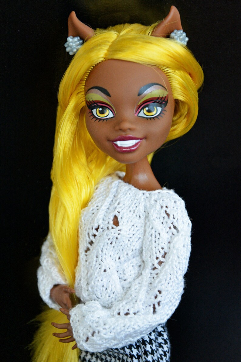May include: A doll with yellow hair, brown skin, and bright makeup. She is wearing a white knitted sweater and a black and white checkered skirt. She has cat ears with silver earrings.