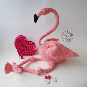 May include: Pink knitted flamingo toy with a long neck and a black beak. The flamingo is lying down with its legs crossed. There is a pink heart-shaped box and two white hearts in the background.