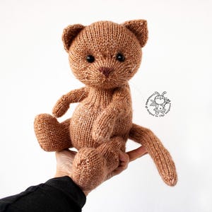Op de afbeelding: Een handgemaakte bruin gebreide kattenpop met zwarte ogen en een lange staart. De kat zit en heeft zijn armen en benen uitgestrekt. De pop wordt in een hand gehouden tegen een witte achtergrond.