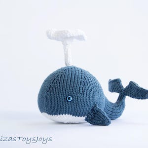 May include: A handmade blue and white knitted whale toy with a white spout. The whale has a small blue eye and a white belly. The tail and fins are also blue. The text "denizasToysjoys" is at the bottom.