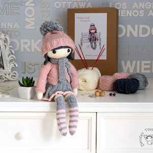 May include: A handmade doll wearing a pink hat, jacket, and striped socks, with a gray skirt and scarf. The doll is part of a knitting kit, with yarn, needles, and a box labeled "Masha Doll Knitting Kit".