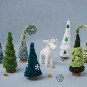 May include: A collection of handmade knitted Christmas tree ornaments in various colors and styles, including green, white, blue, and brown. The ornaments are decorated with small green felt leaves and a white felt snowflake. A white ceramic moose figurine is also included in the image.