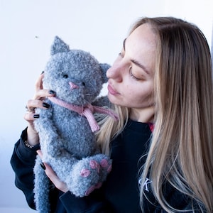 May include: A handmade gray plush cat with pink accents, including a nose, paw pads, and a scarf. The cat is held in front of a white background.