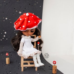 Könnte beinhalten: Eine Puppe in einem weißen Kleid und einem roten Pilz-Hut sitzt auf einem Holzstuhl. Die Puppe hat langes braunes Haar und hält einen roten und goldenen Pilz-Anhänger.