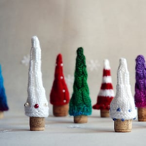 Puede incluir: Una colección de diez árboles de Navidad en miniatura tejidos a mano en varios colores, incluyendo rojo, blanco, verde, azul y morado. Los árboles están decorados con cuentas blancas y azules y se muestran en bases de corcho.