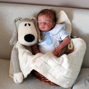 May include: A knitted gray and white dog toy is nestled in a white knitted blanket with a baby doll wearing a blue and white sailor shirt. The blanket is in a wicker basket.