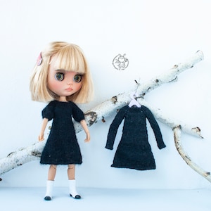 May include: A doll wearing a black knitted dress with puffed sleeves and white boots. A second black knitted dress hangs on a branch behind the doll.