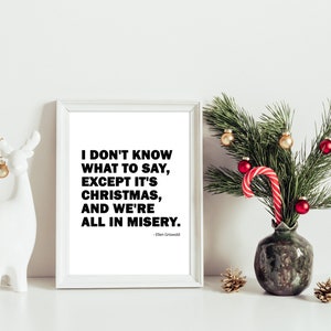 Puede incluir: Una impresión enmarcada en blanco con una cita en blanco y negro de la película Vacaciones de Navidad. La cita dice "No sé qué decir, excepto que es Navidad, y estamos todos en la miseria." - Ellen Griswold. La impresión está sobre una superficie blanca con una figurita de reno de cerámica blanca y un jarrón verde con una rama de pino y adornos rojos y dorados.