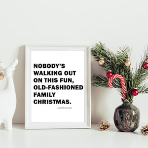 May include: A white framed print with a black and white quote from the film "Christmas Vacation" that reads "Nobody's walking out on this fun, old-fashioned family Christmas." The quote is attributed to Clark W. Griswold. The print is sitting on a white surface with a white ceramic reindeer figurine and a green vase with a pine branch and red and gold baubles.