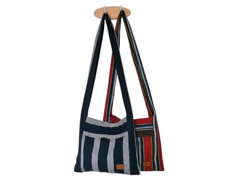May include: Two shoulder bags with long straps hang from a wooden fixture. One bag is navy blue with white vertical stripes, and the other is red with blue and green stripes. Both bags have a rectangular shape.