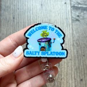 May include: A badge reel featuring a glossy, arched topper with a light blue background and the text WELCOME TO THE SALTY SPLATOON in bold blue letters. The centre displays an illustration of a building with a sign reading THE SALTY SPITOON.